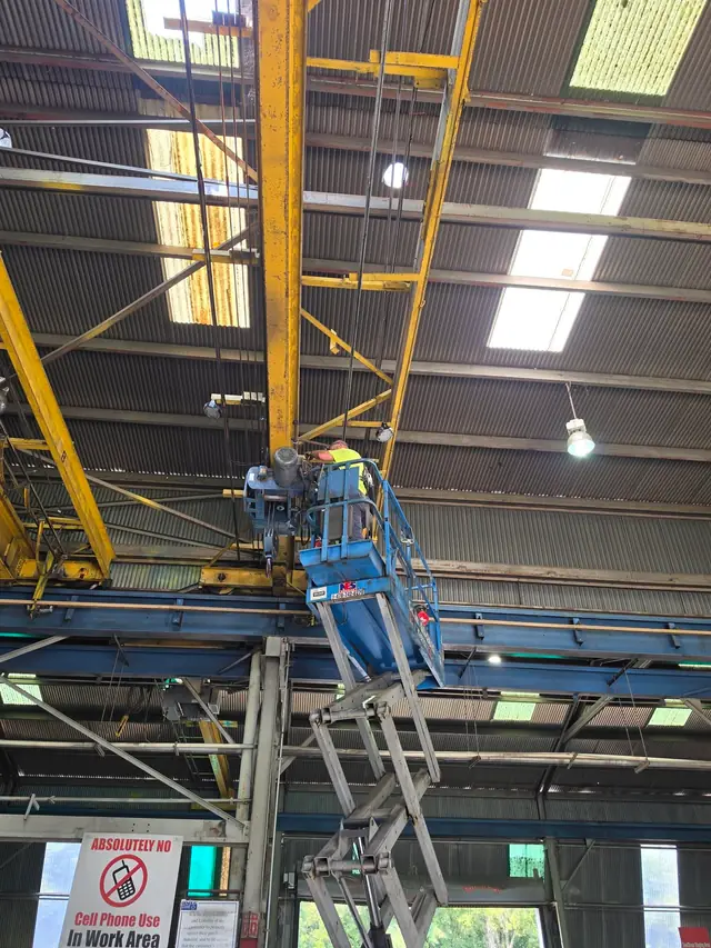 4A Electrical crew member in hi-vis on a blue boom lift beside a yellow overhead crane and high-bay lights