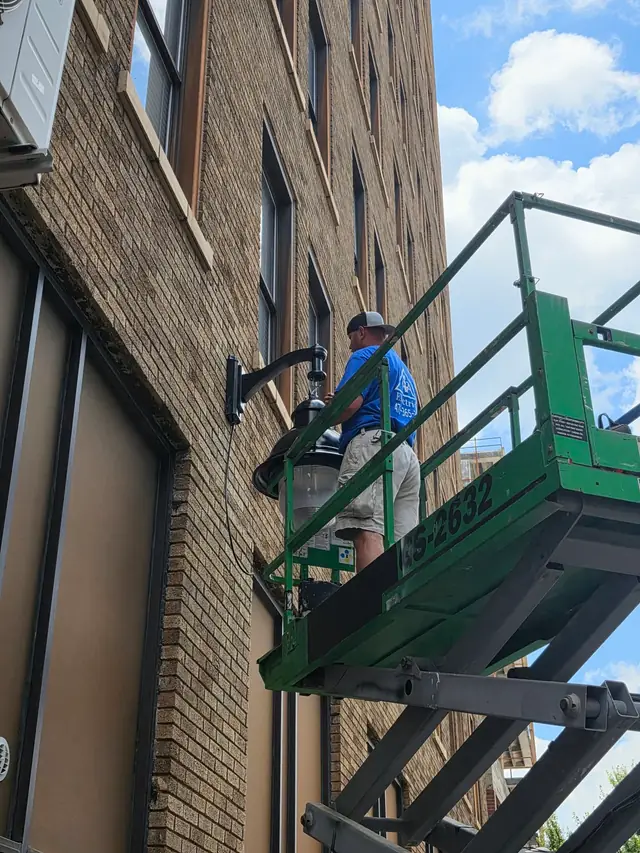 4A Electrical crew member in a branded blue shirt on a green scissor lift fitting a gooseneck lantern to brick